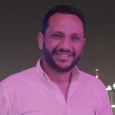 Smiling young woman with curly hair sitting at a laptop, in front of a teal wall.